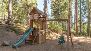 A wooden playhouse with a slide and swings in a forested area. - Greene Gables- Cozy Mountain Retreat (Avery)