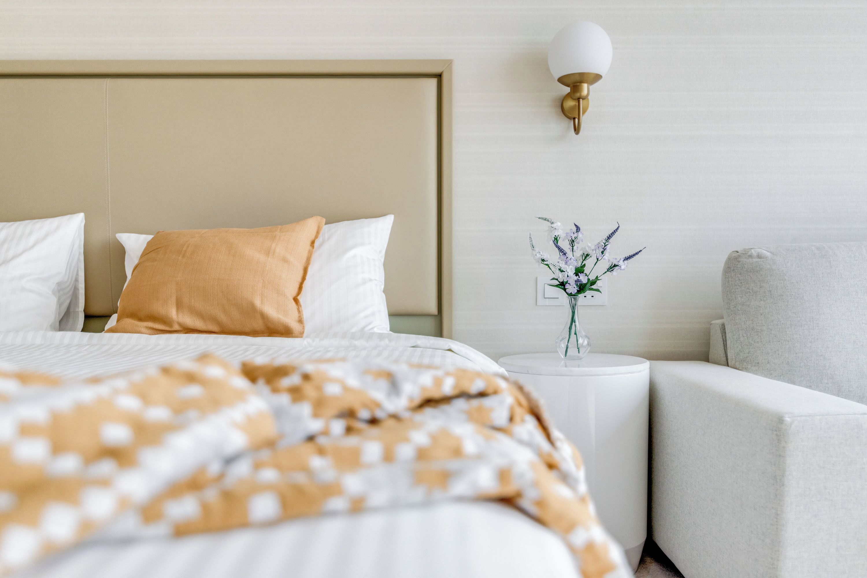 Ein ordentlich bezogenes Bett mit einem gemusterten Überwurf, ein Nachttisch mit Blumen in einer Vase und ein Sessel in einem modernen Hotelzimmer.