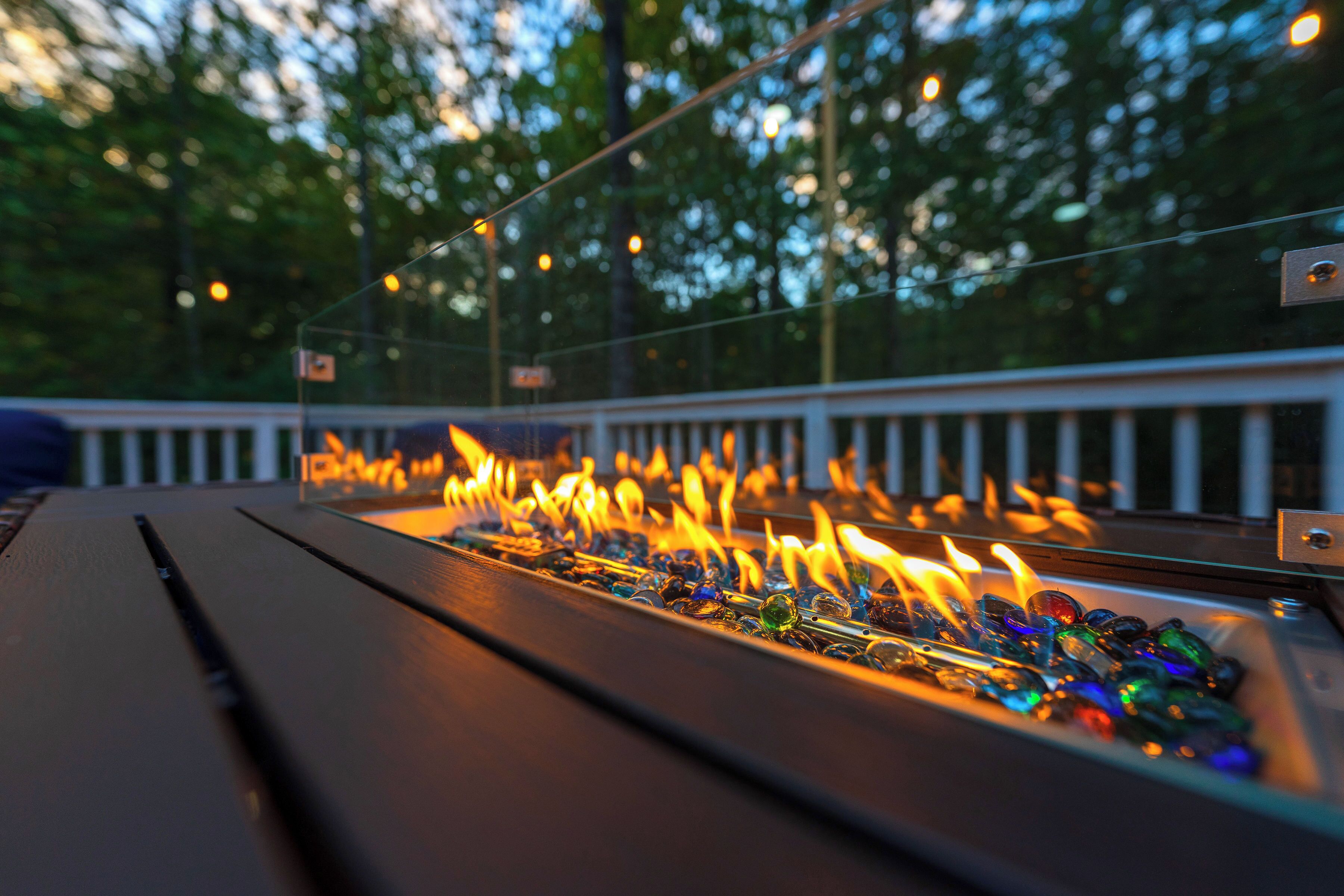 Ein Tisch mit Glasplatte im Freien, ausgestattet mit einem Feuerkorb, umgeben von einem Geländer und Bäumen im Hintergrund.