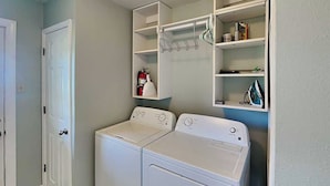 A laundry room with two white front-loading washers and dryers, a fire extinguisher on the wall, and shelves above holding various items.