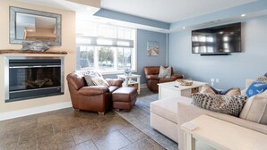 A modern living room with leather sofas, a fireplace, a flat-screen TV, and a large window providing natural light. - Stunning Downtown Condo w/ Water Views (Grand Haven)
