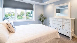 A bedroom with a large bed, a dresser, a mirror, and a window with blinds. - Stunning Downtown Condo w/ Water Views (Grand Haven)