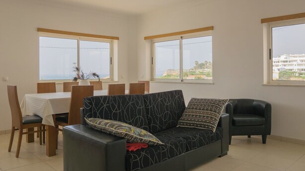 Uma sala de estar com um sofá, uma mesa de jantar com cadeiras e amplas janelas com vista para edifícios e vegetação.