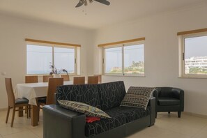A living room with a sofa, a dining table with chairs, and large windows offering a view of buildings and greenery.