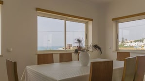 Uma área de refeições com uma mesa posta para quatro pessoas, cadeiras de madeira, toalha de mesa branca e um vaso com plantas secas.
