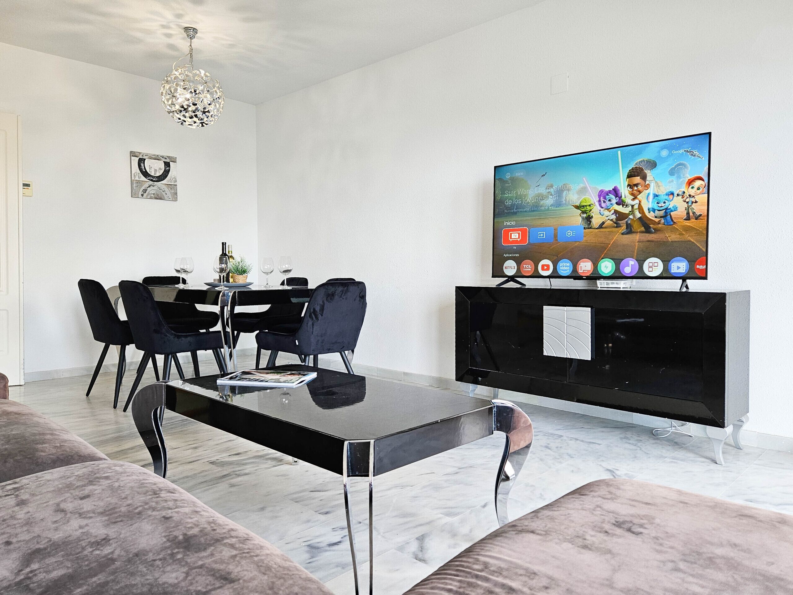 A modern living room with a dining area, a flat-screen TV, and a chandelier.
