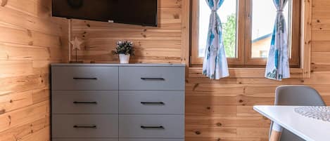 Interior de una cabaña de madera con un aparador gris, un televisor de pantalla plana montado encima y una ventana con cortinas florales.