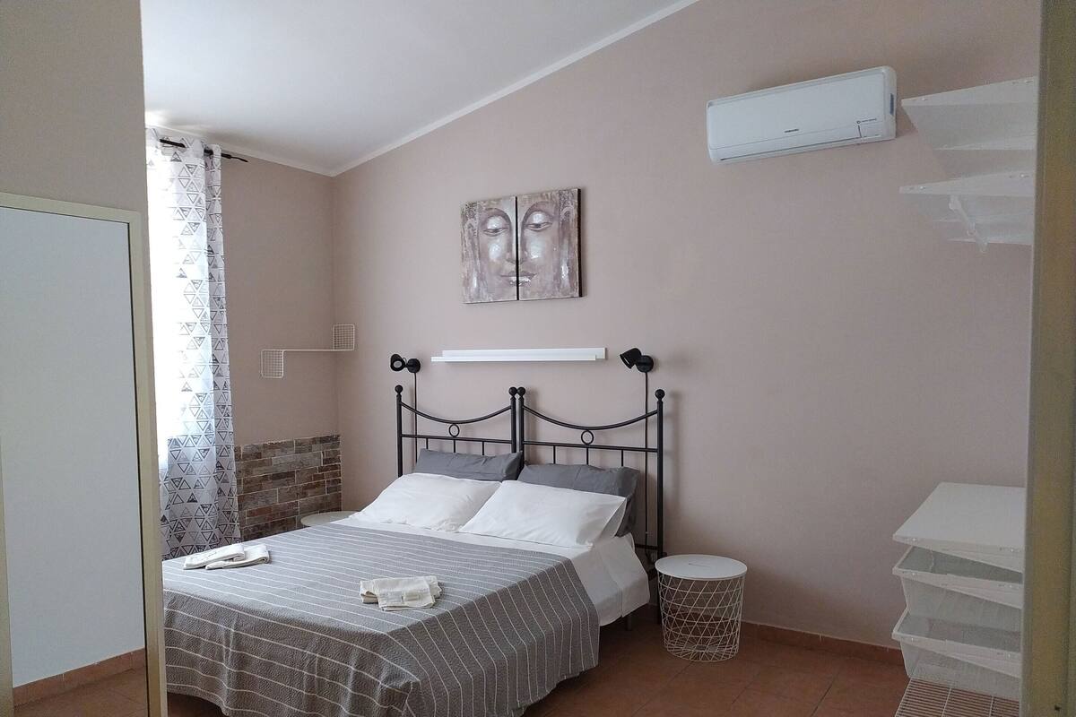 Un dormitori amb un llit, aire condicionat fixat a la paret, una taula rodona i petita, i una estant amb una fotografia.