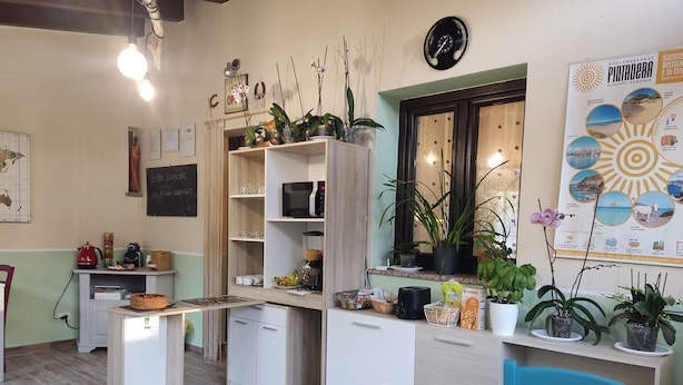 A compact kitchen with a dining area, equipped with a microwave, kettle, and various kitchen utensils. There are potted plants and a window with a view. - Guesthouse Pintadera (Sassari)