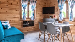 Interior de una cabaña de madera con sofá, mesa de comedor, sillas, televisor y cortinas.