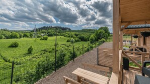 Un'elegante terrazza in legno con una panchina e cesti fioriti, affacciata su un prato recintato con linee elettriche e alberi sullo sfondo.