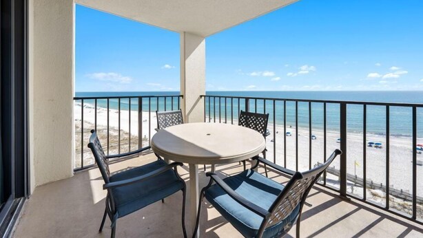 A balcony with a round table and four chairs, overlooking a beach with a clear view of the ocean and beachgoers.