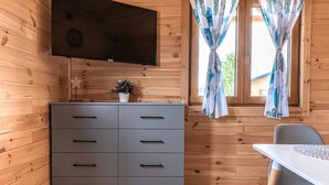 A wooden cabin interior with a grey dresser, a flat-screen TV mounted above it, and a window with floral curtains. - Cottage in Duszniki Zdrój with Hilltop Views (Duszniki Zdrój)