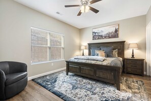 A bedroom with a bed, a chair, a nightstand, a lamp, a ceiling fan, and a window with blinds. - Branson’s Like Home Hideaway (Branson)