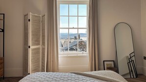 A bedroom with a large window, a bed with a textured blanket, a wooden wardrobe, and a mirror on the wall.