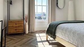 A bedroom with a bed, a dresser, a vase with dried plants, and a window with a view of buildings.