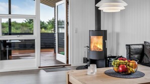 Ein modernes Wohnzimmer mit Kaminofen, einem Esstisch mit Obstschale und Blick durch eine Glastür auf eine Terrassenanlage.