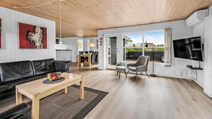 Ein modernes Wohnzimmer mit Holzboden, einem schwarzen Ledersofa, einem Holztisch mit Obst, einem grauen Sessel und einem Fernseher.