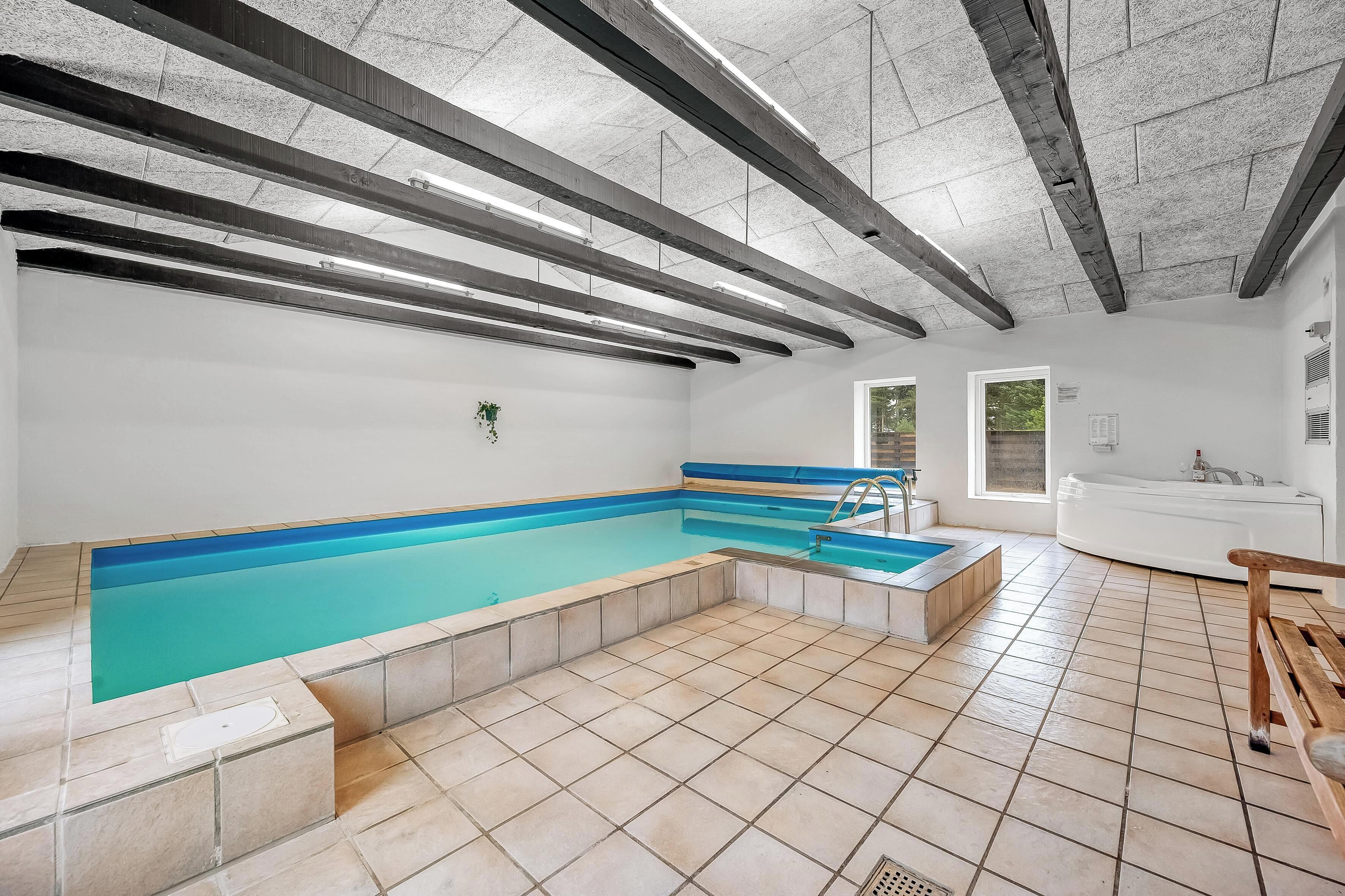 Una piscina con bordo blu, una panca di legno e una vasca da bagno bianca in una stanza con pavimento piastrellato e soffitto in cemento.