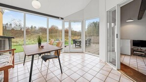 Un patio amplio con una mesa y sillas de comedor, puertas correderas de vidrio que dan a un jardín, y un televisor empotrado.