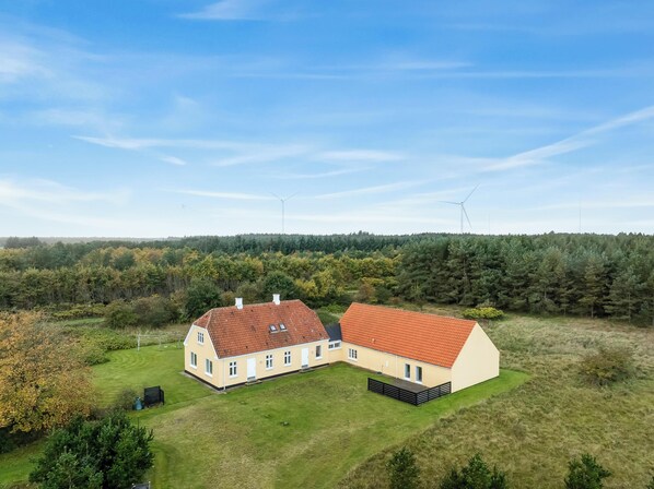 A two-story building with a red-tiled roof, surrounded by a grassy area and dense forest. - Large pool house with activity room located in peaceful surroundings. (Frøstrup)