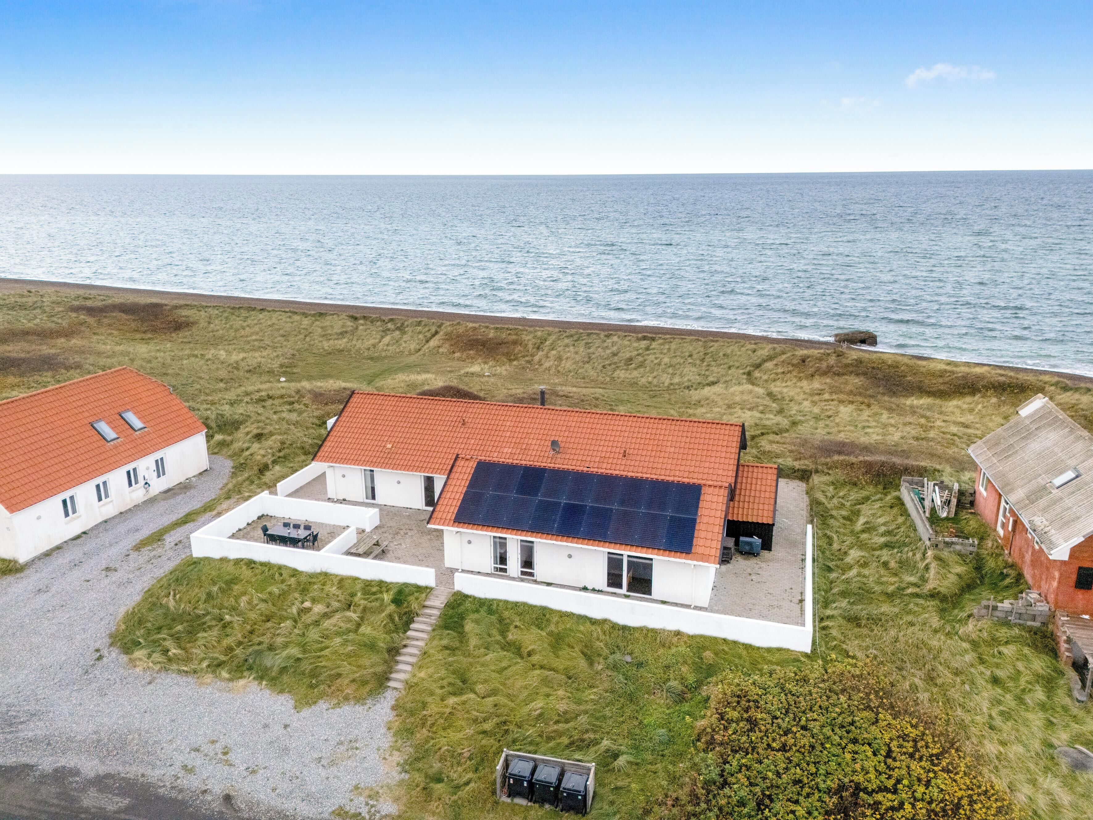 A coastal property with a main house featuring a red-tiled roof and solar panels, surrounded by grassy fields and a view of the sea.