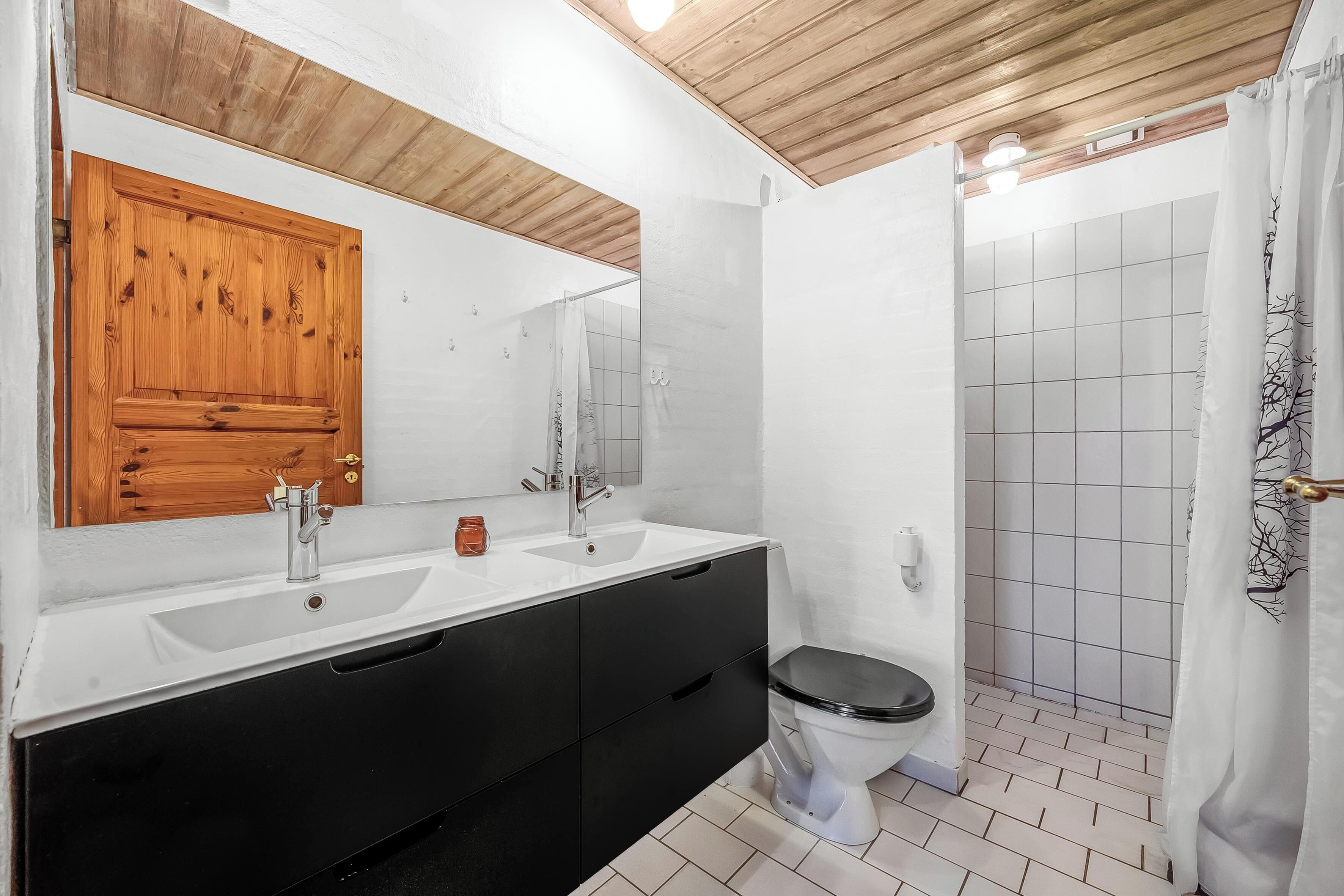 A bathroom with a wooden ceiling, a white sink, a black cabinet, a toilet, and a shower area.