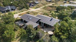 A modern house with a large deck and a hot tub, surrounded by a natural landscape and other houses.