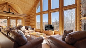 L’intérieur d’une cabane en rondins, avec une grande fenêtre, des canapés en cuir, un téléviseur et une cheminée en pierre.