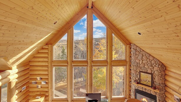 L’intérieur d’une cabane en rondins, avec une cheminée en pierre, de grandes fenêtres et un ventilateur de plafond.