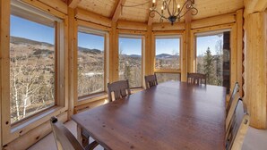 Une salle à manger en bois, avec une grande table et des chaises, de grandes fenêtres offrant une vue sur un paysage montagneux.