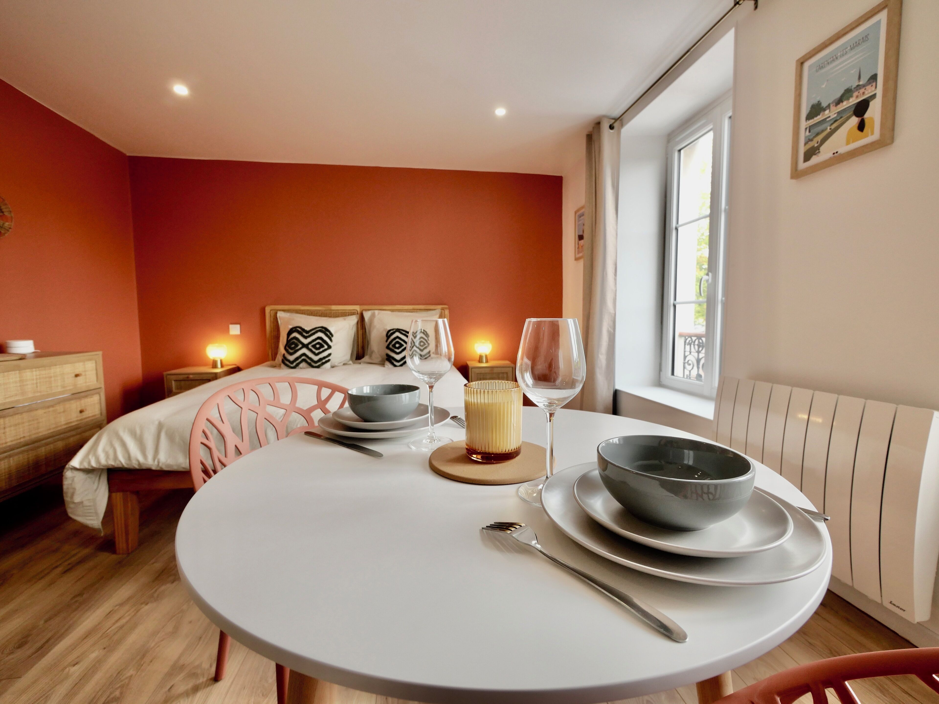 A dining area with a round table set for two, a bed with patterned pillows, and a window with a view.