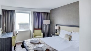 Una habitación de hotel con cama, escritorio, silla, lámpara y ventana con cortinas.