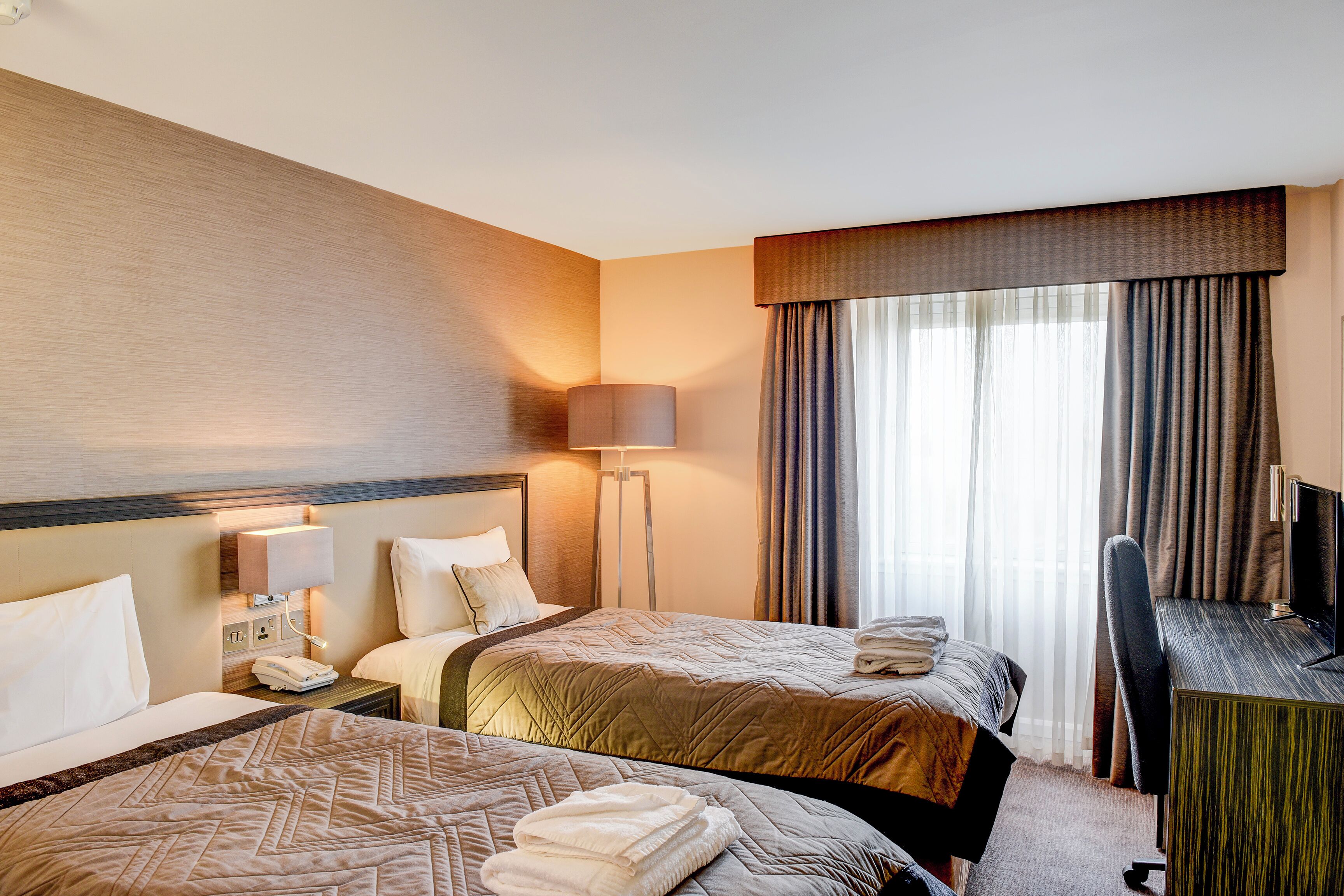 A hotel room with two beds, a desk, a lamp, and a large window with curtains.