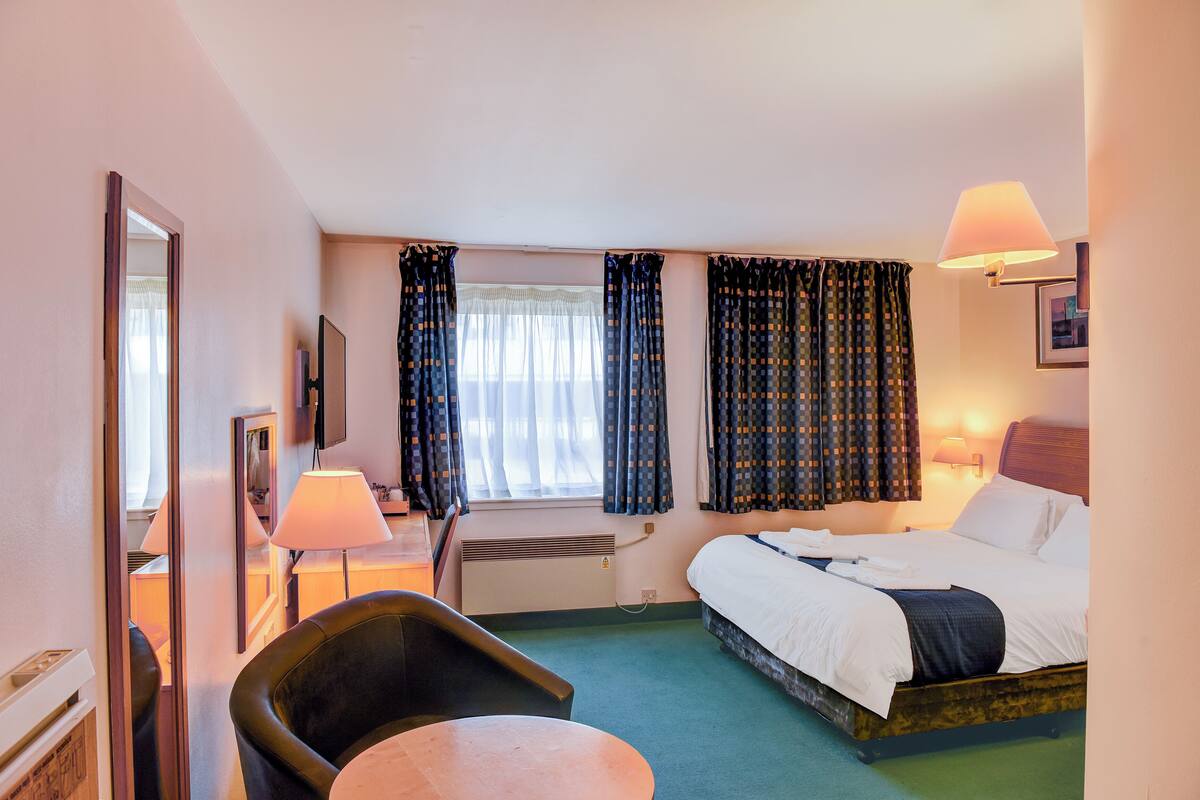 A hotel room with a bed, a chair, a table, a mirror, a TV, and a window with curtains.