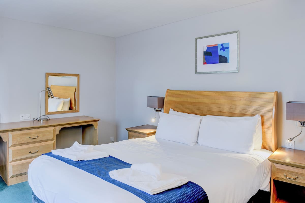 A hotel room with a wooden bed, a bedside table, a nightstand, a mirror, and a framed artwork on the wall.