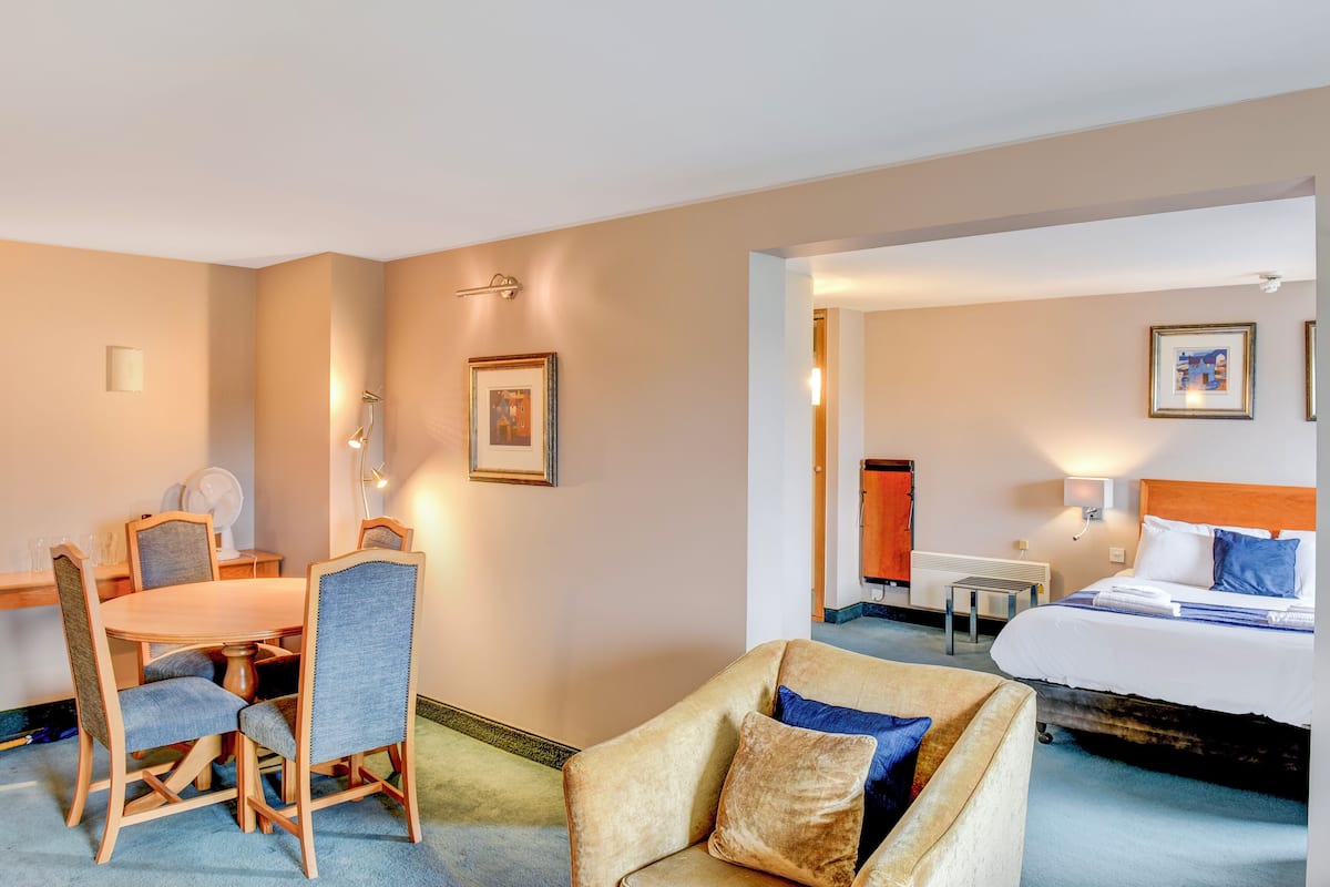 A hotel room with a bed, a chair, a dining table with chairs, a small table, and a picture on the wall.