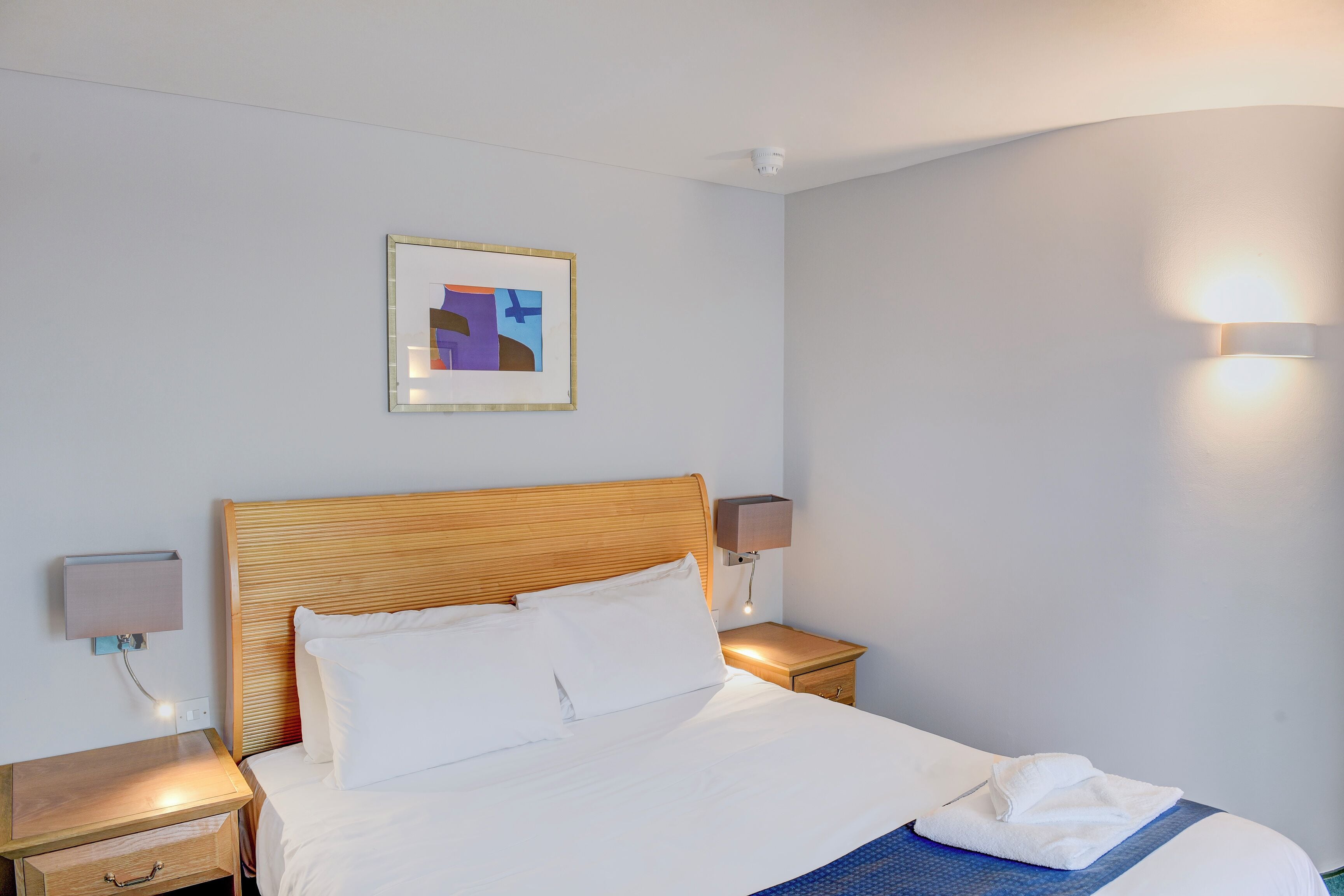 A neatly made bed with white linens, a wooden headboard, and a blue accent on the bedspread. There is a framed abstract artwork on the wall above the bed and a wall-mounted lamp beside the bed.