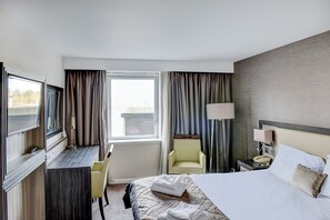 Une chambre d’hôtel comprenant un lit, un bureau avec une chaise, une télévision, une lampe et une fenêtre avec des rideaux.
