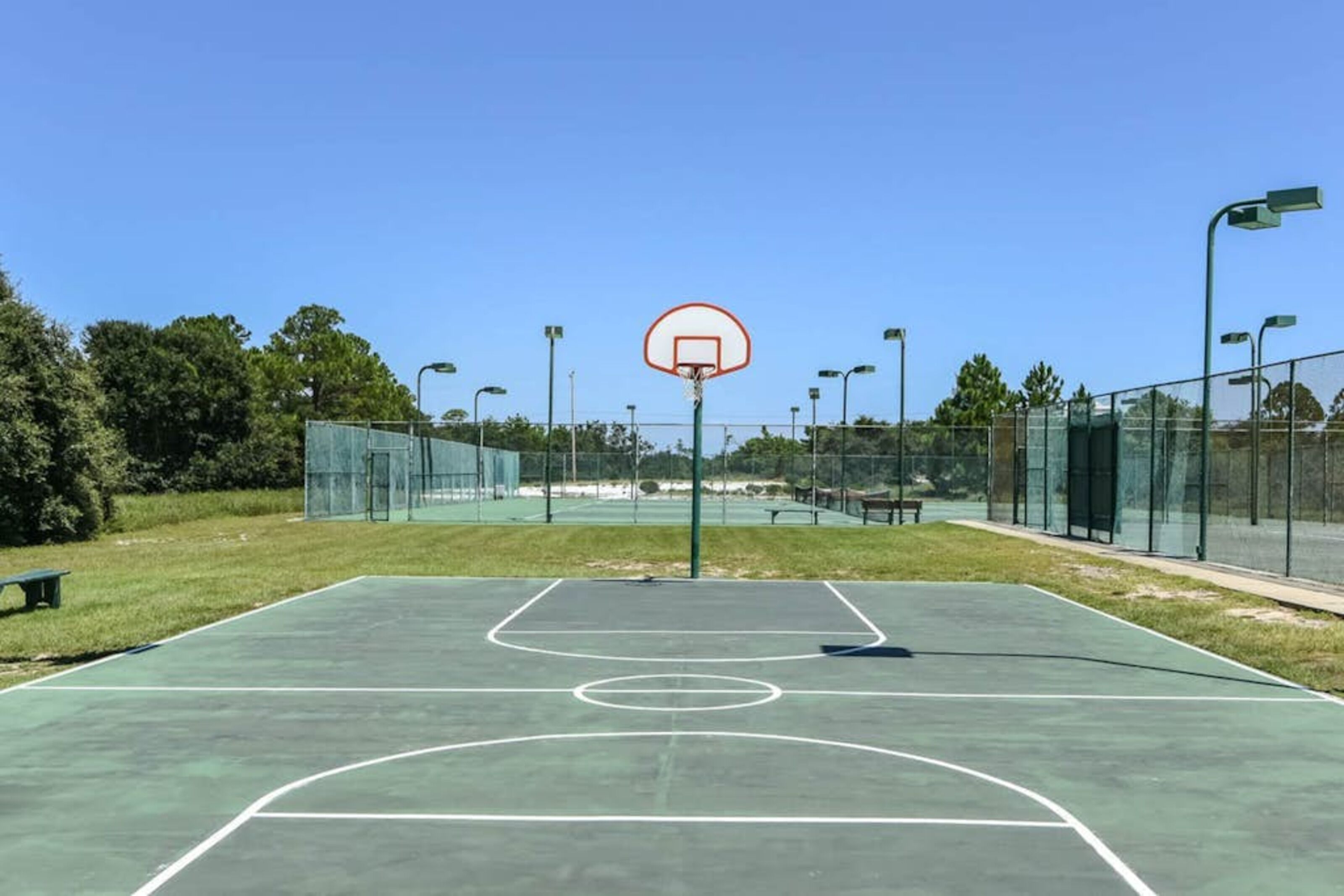 An outdoor basketball court with a hoop, surrounded by a fence and trees.