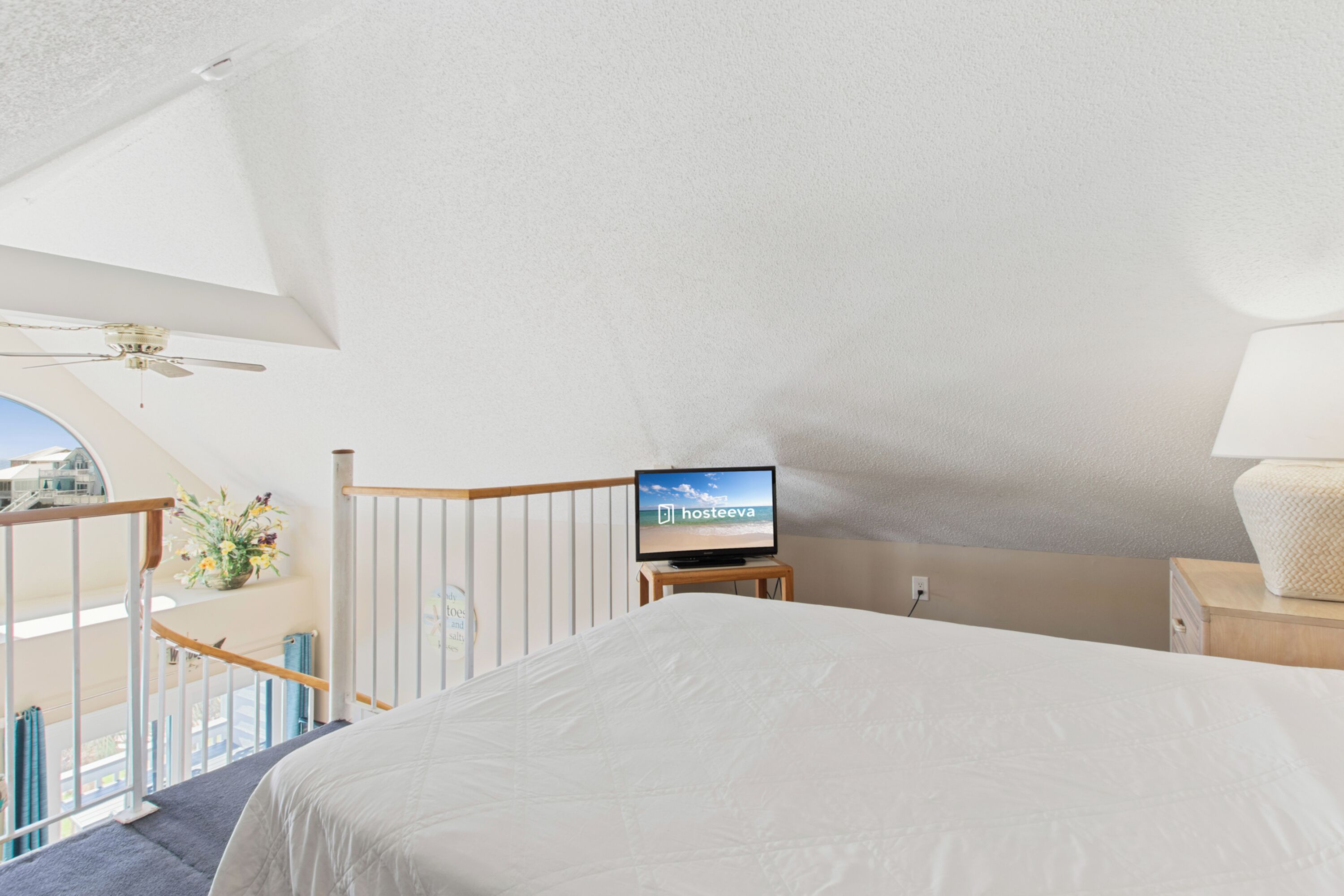Un dormitorio con cama, televisor sobre un mueble, mesita de noche, lámpara, ventilador de techo y balcón con barandilla.