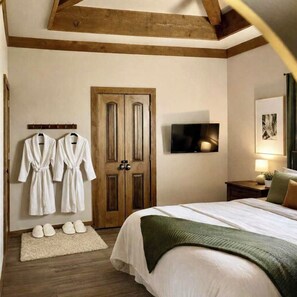 Habitación de hotel con puerta de madera, cama con una manta verde, televisor en la pared y dos batas blancas colgadas en un perchero.