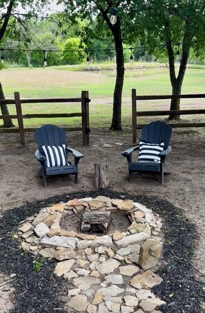 Deux fauteuils Adirondack avec des coussins à rayures, un foyer en pierre et une clôture en bois, le tout dans un cadre naturel.