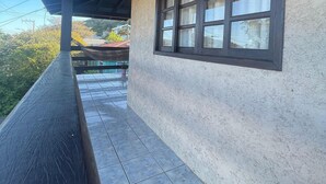 A covered balcony with a tiled floor, a window with multiple panes, and a view of greenery and buildings in the background. - Oásis Morro das Pedras - Floripa (Florianópolis)