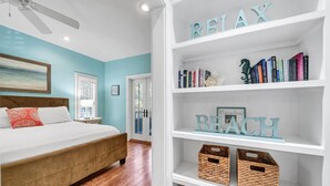 Un dormitorio con cama, ventilador de techo, estantería con libros y objetos decorativos, y vista al exterior.