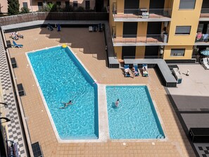 Una piscina en la azotea con dos zonas separadas, rodeada de sillones de descanso y un edificio con balcones.