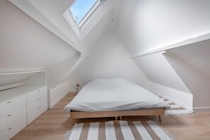 Una habitación pequeña con techo inclinado, una cama, un tocador blanco y un tragaluz.