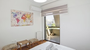 A bedroom with a bed, a small table, a bench, an air conditioning unit, a radiator, and a large window with a view of the outside. - Afrodite Classic Residence, 2BR, WiFi, City View (Paleo Faliro)