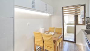 A compact kitchen with white cabinets, a dining table with yellow chairs, and a sliding glass door leading to a balcony. - Afrodite Classic Residence, 2BR, WiFi, City View (Paleo Faliro)