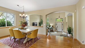 A dining area with a white table, yellow chairs, and a chandelier. There is a large window with a view of trees and a door leading to another room. - 1/2 Acre Retreat: 100" TV, Open layout Navy Base and Six Flags Great America (Gurnee)